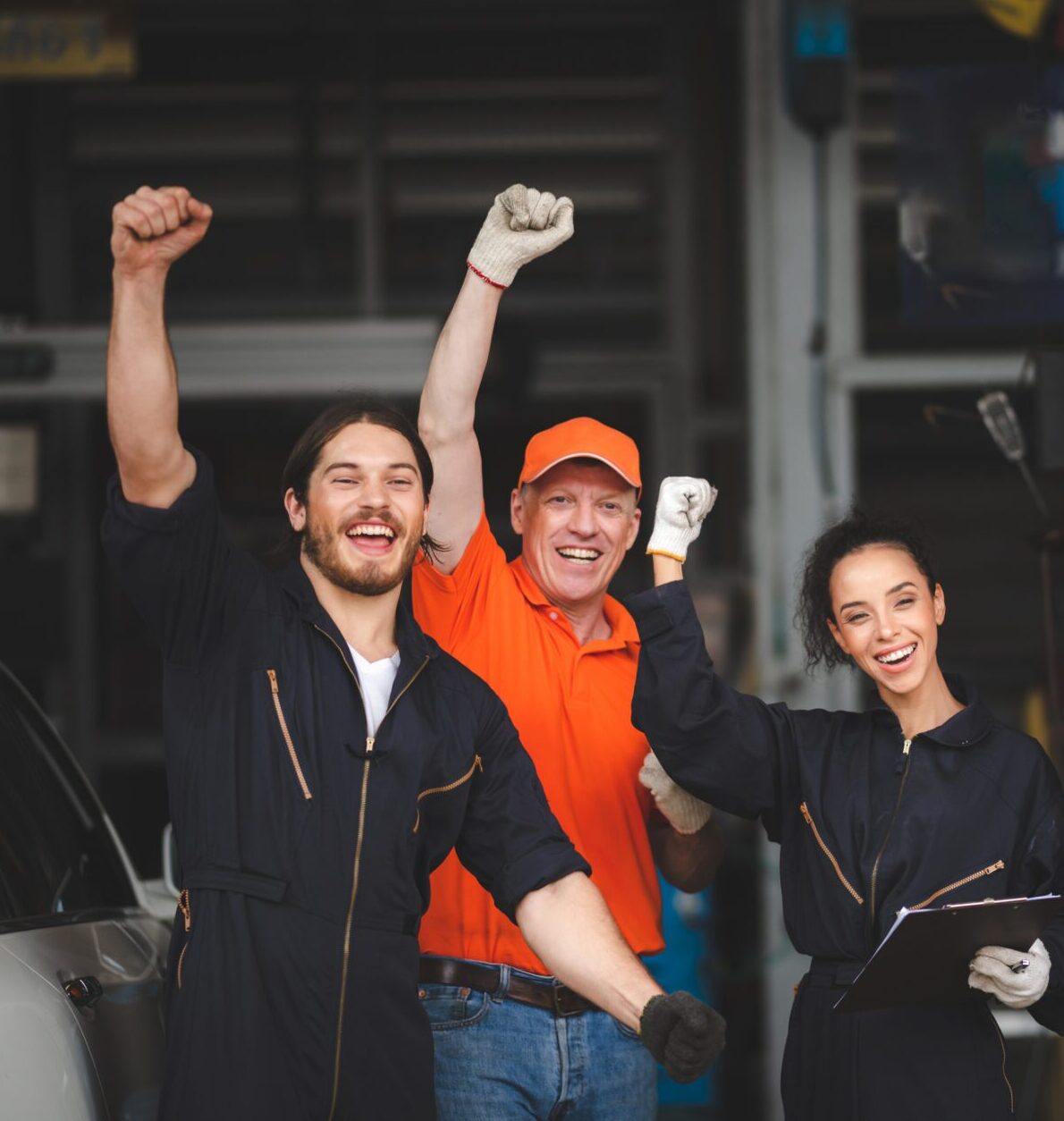 Group of young and senior male and female car mechanics in garage wearing uniform enjoying and celebrating with female worker holding clipping board